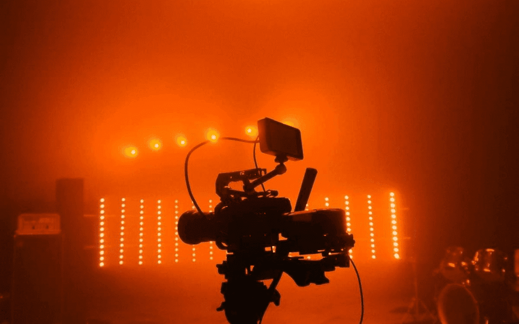 Camera filming a live performance under dramatic orange stage lighting.