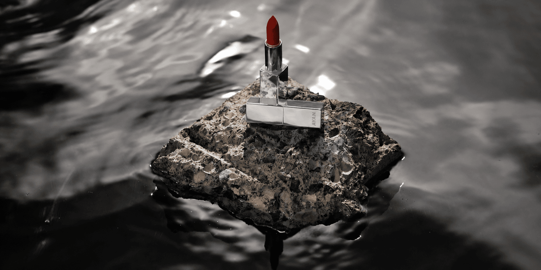 Ayan Beauty lipstick displayed on a textured rock surrounded by reflective water